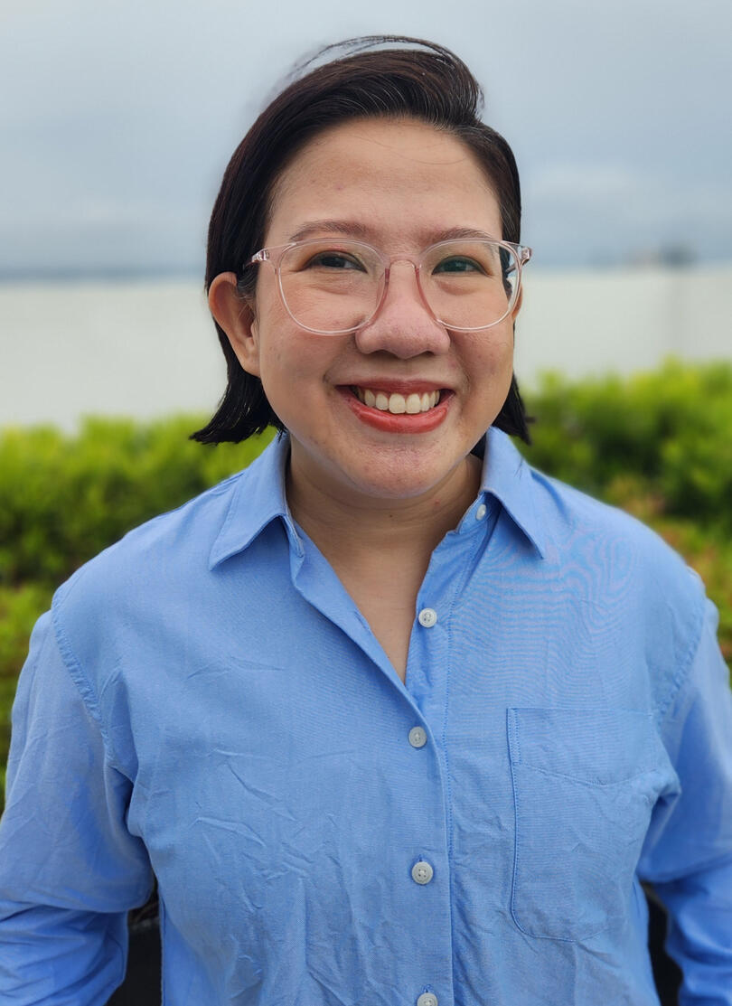 Luna Javier A Filipino woman with mid-length dark hair and glasses, wearing a striped sweater and jeans, smiles at the camera as she sits on a daybed.